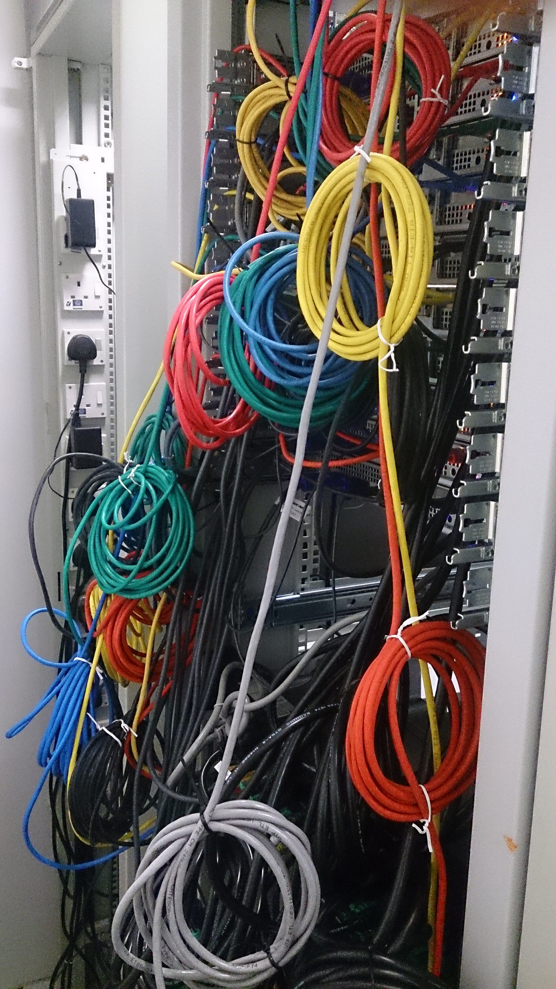 A server rack crowded with dozens of coiled, color-coded network cables tied with zip ties, and a power strip on the left.