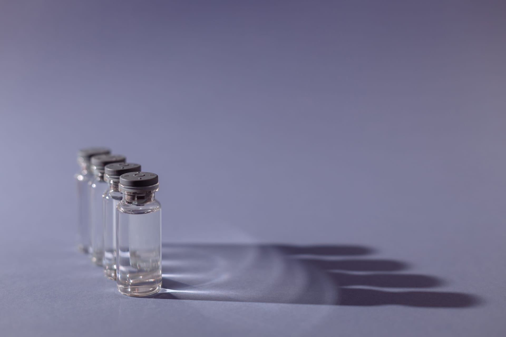 Cuatro frascos de vidrio transparente con tapa gris, alineados sobre una superficie violeta, proyectan sombras largas hacia la derecha.