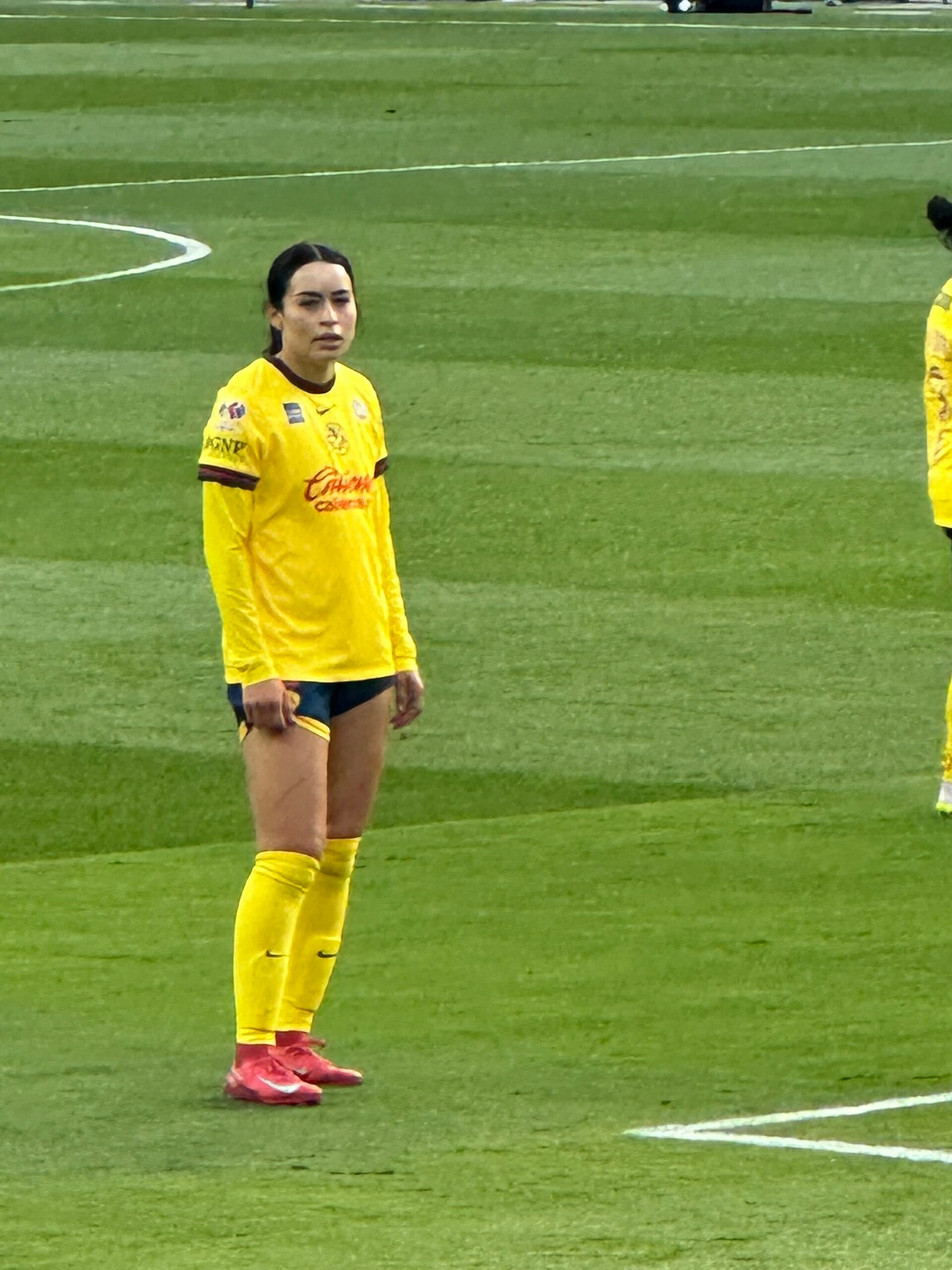 Una futbolista de camiseta amarilla de manga larga y calcetas amarillas espera de pie en el césped durante un partido.