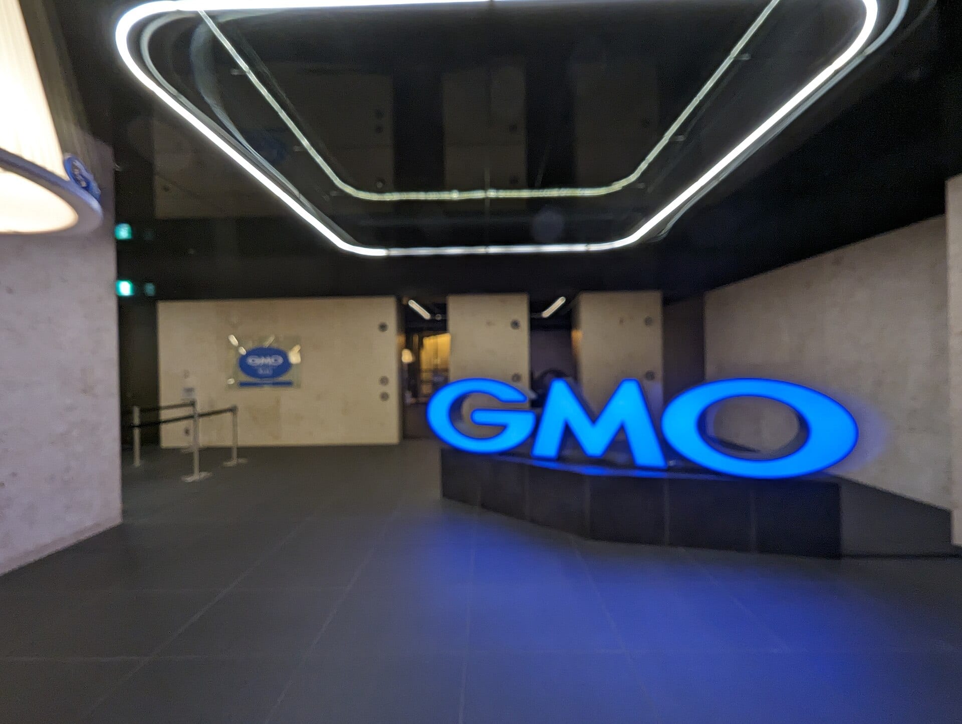 El logotipo azul iluminado de GMO sobre una base oscura en un vestíbulo de paredes de concreto.