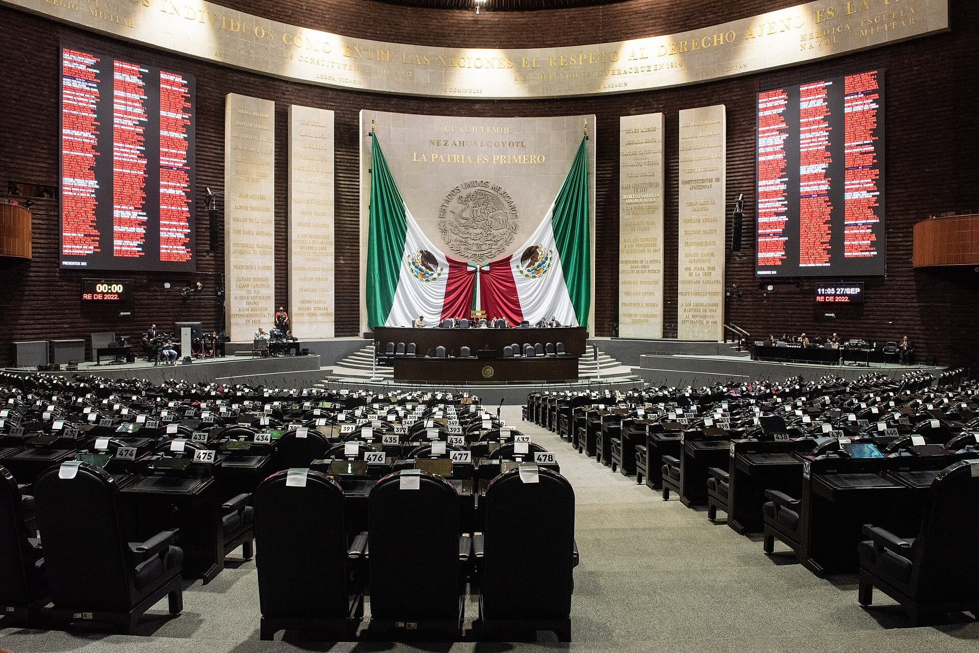Salón de sesiones de la Cámara de Diputados visto desde las curules vacías, con una bandera de México tras la mesa directiva y tableros de votación a los lados.