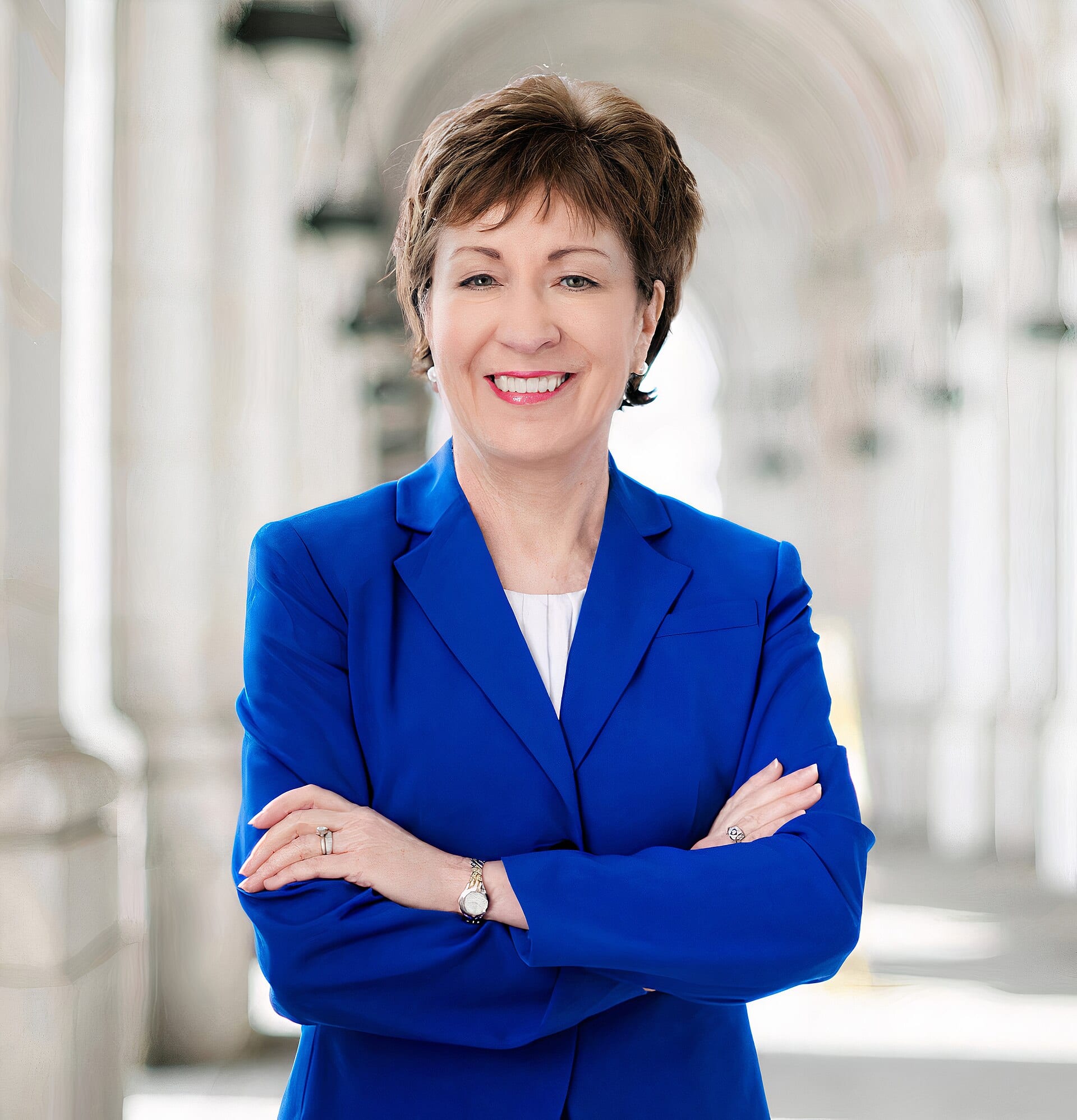 La senadora Susan Collins, de saco azul y blusa blanca, sonríe con los brazos cruzados en un pasillo con arcos.
