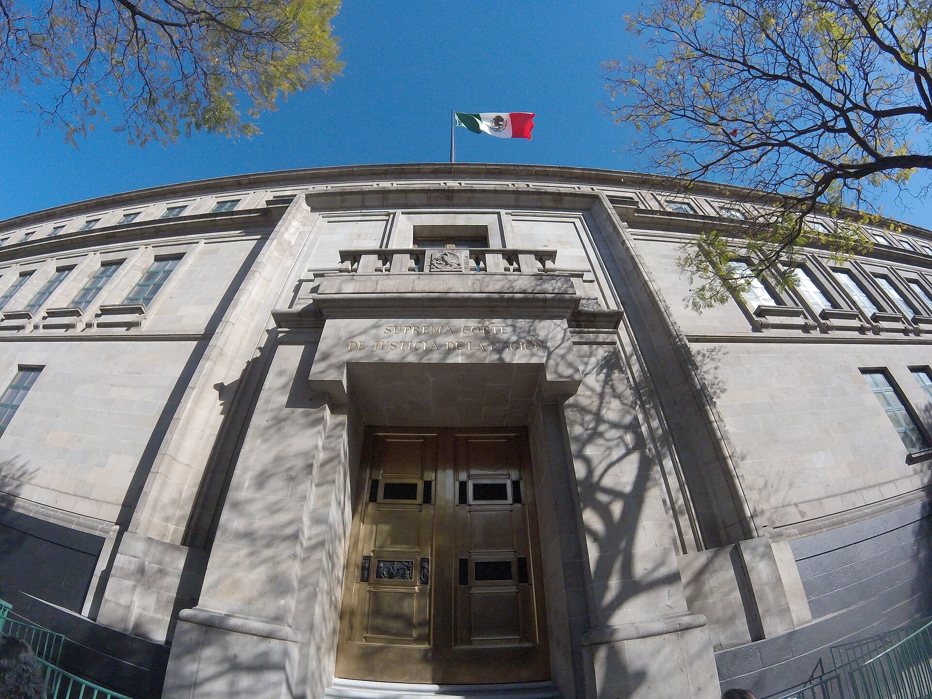Fachada en contrapicado de la Suprema Corte, con la bandera de México ondeando sobre el pórtico de piedra y las puertas de bronce.