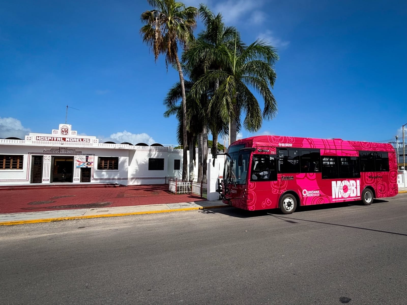 Un autobús eléctrico rosa con la marca MOBI estacionado frente a un edificio blanco, junto a unas palmas.