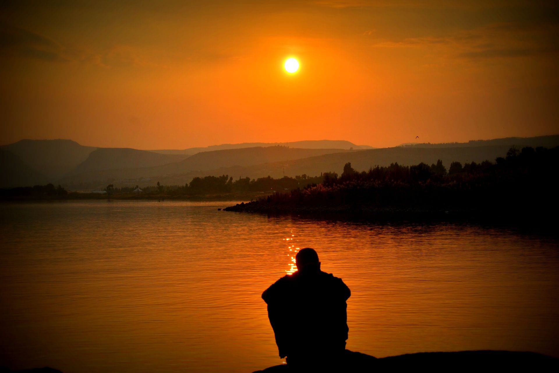 La silueta de una persona sentada de espaldas frente a un lago, con el sol bajo un cielo naranja y colinas al fondo.