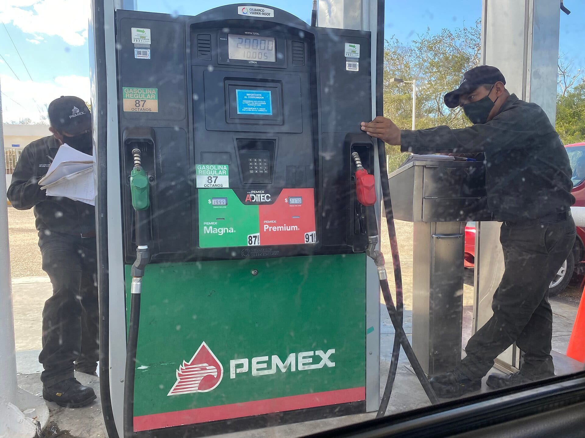 Una bomba de gasolina Pemex con sus mangueras verde y roja y dos trabajadores con cubrebocas a los lados.