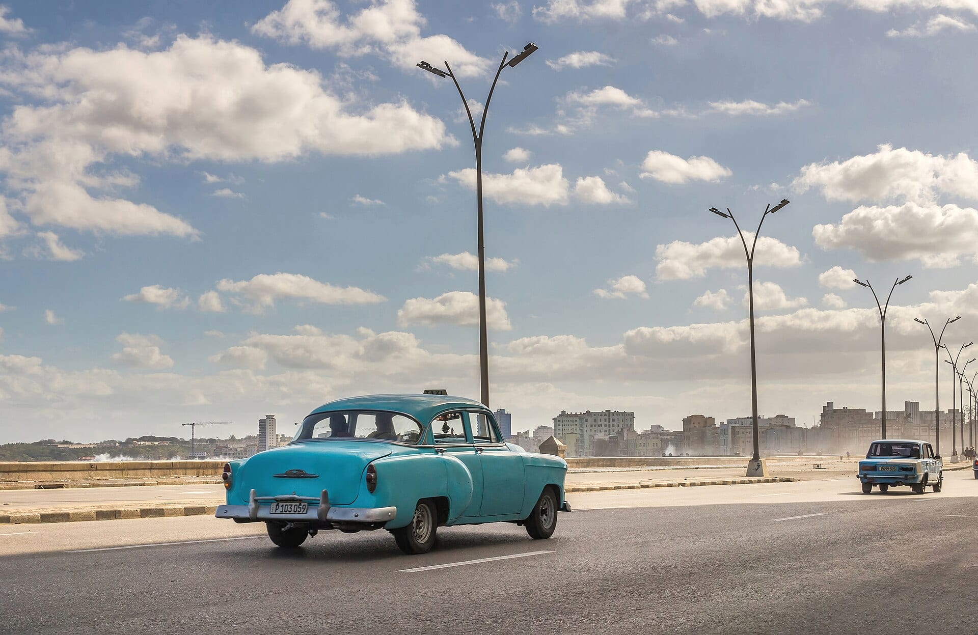 Un taxi clásico color turquesa circula por el Malecón de La Habana, con edificios al fondo.