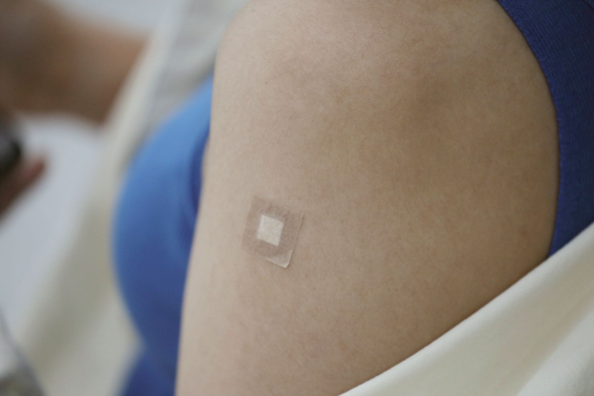 Un brazo con un parche adhesivo cuadrado sobre la piel, junto a una manga azul y una bata blanca desenfocadas.