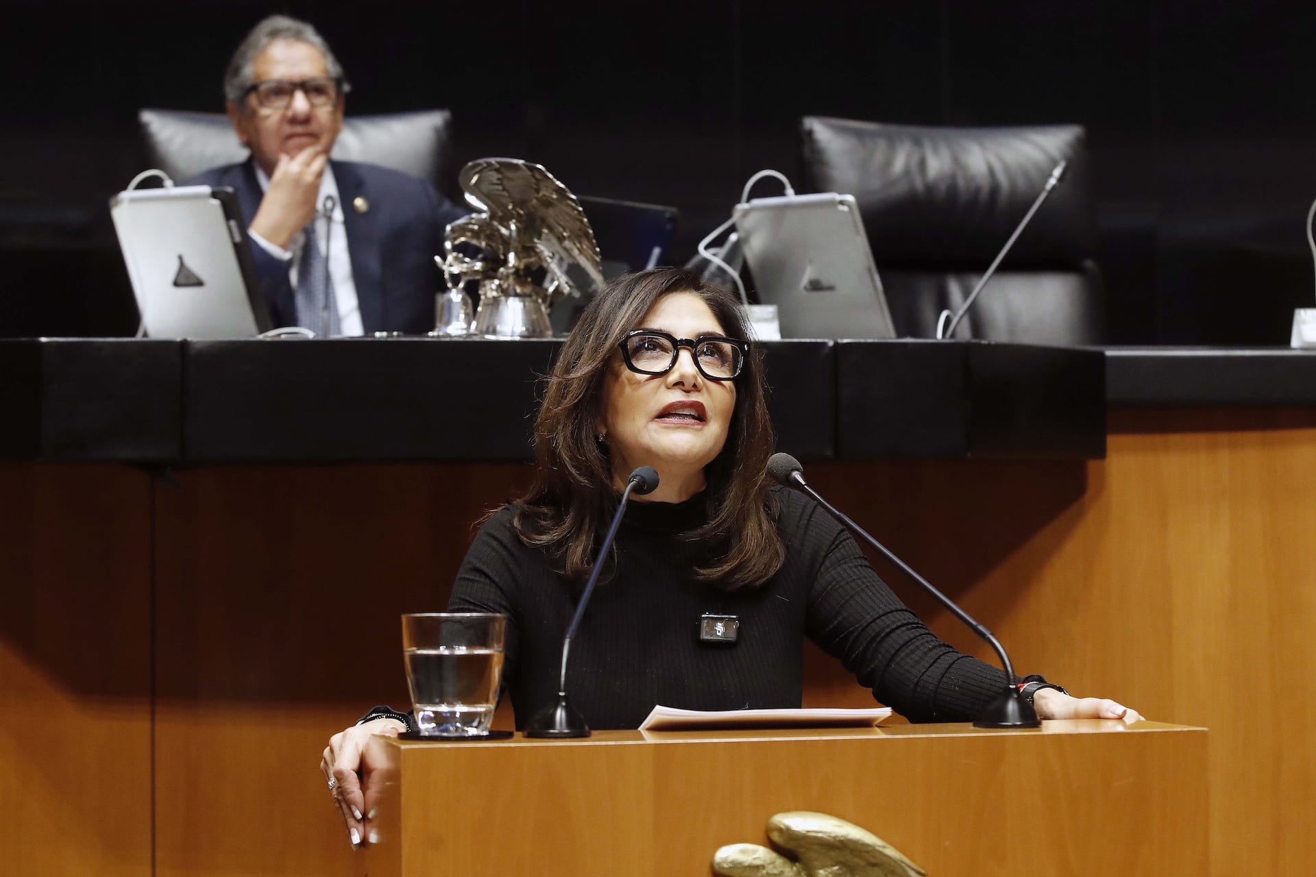 La senadora Alejandra Barrales habla desde el atril de la tribuna del Senado, con un legislador sentado detrás en la mesa directiva.