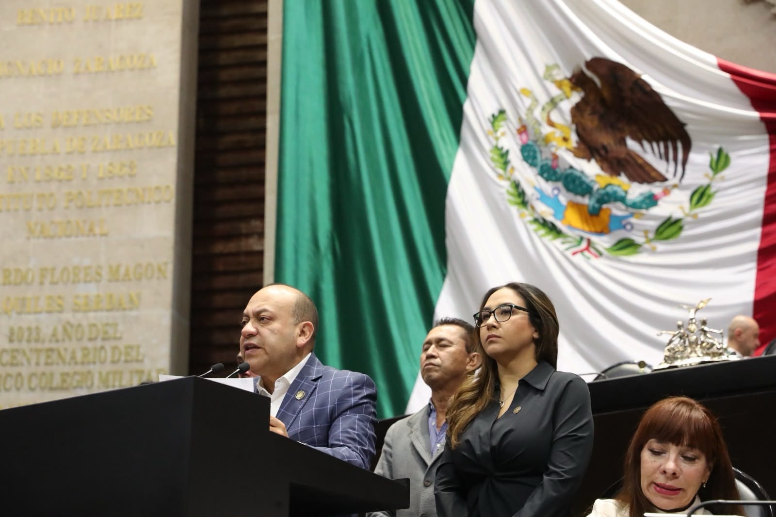 Un diputado habla desde la tribuna del Pleno de San Lázaro; detrás de él, la bandera de México y dos legisladoras.