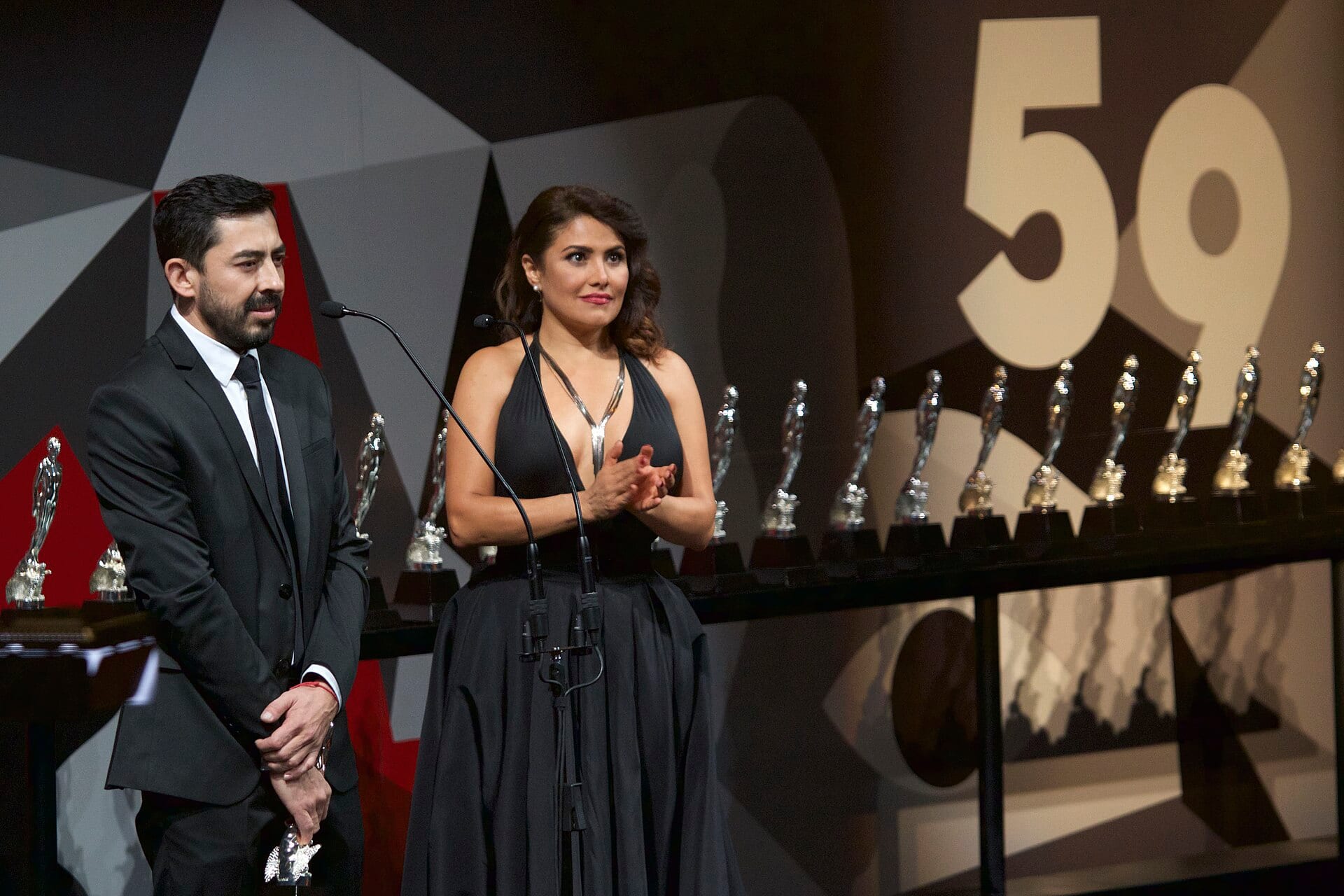 A man in a suit and a woman in a black gown on stage at the 59th Ariel Awards ceremony in 2017, in front of a row of statuettes.