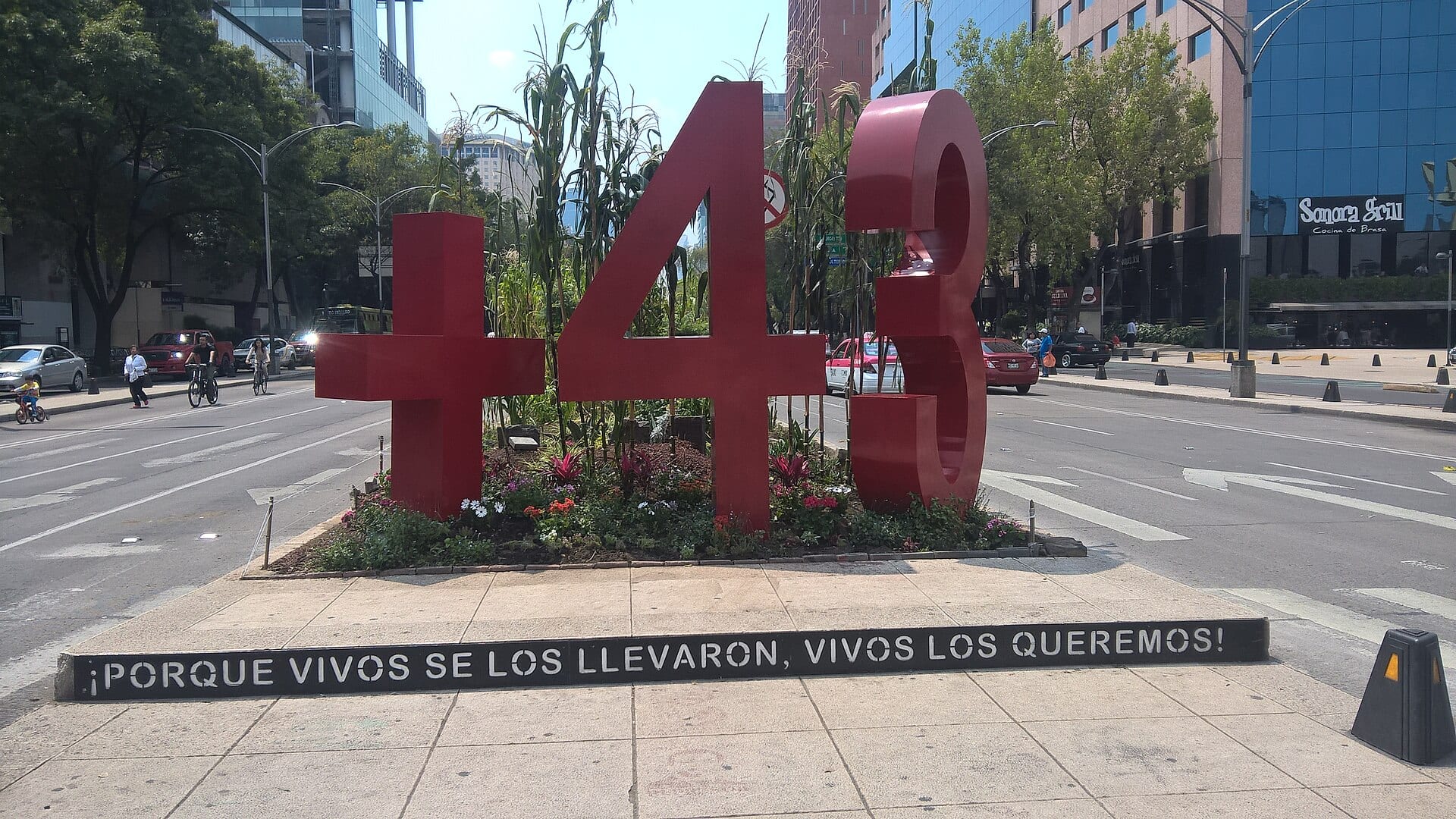 Escultura roja con el signo más, el número 43 y una franja con la leyenda "Porque vivos se los llevaron, vivos los queremos", sobre un camellón de Paseo de la Reforma.
