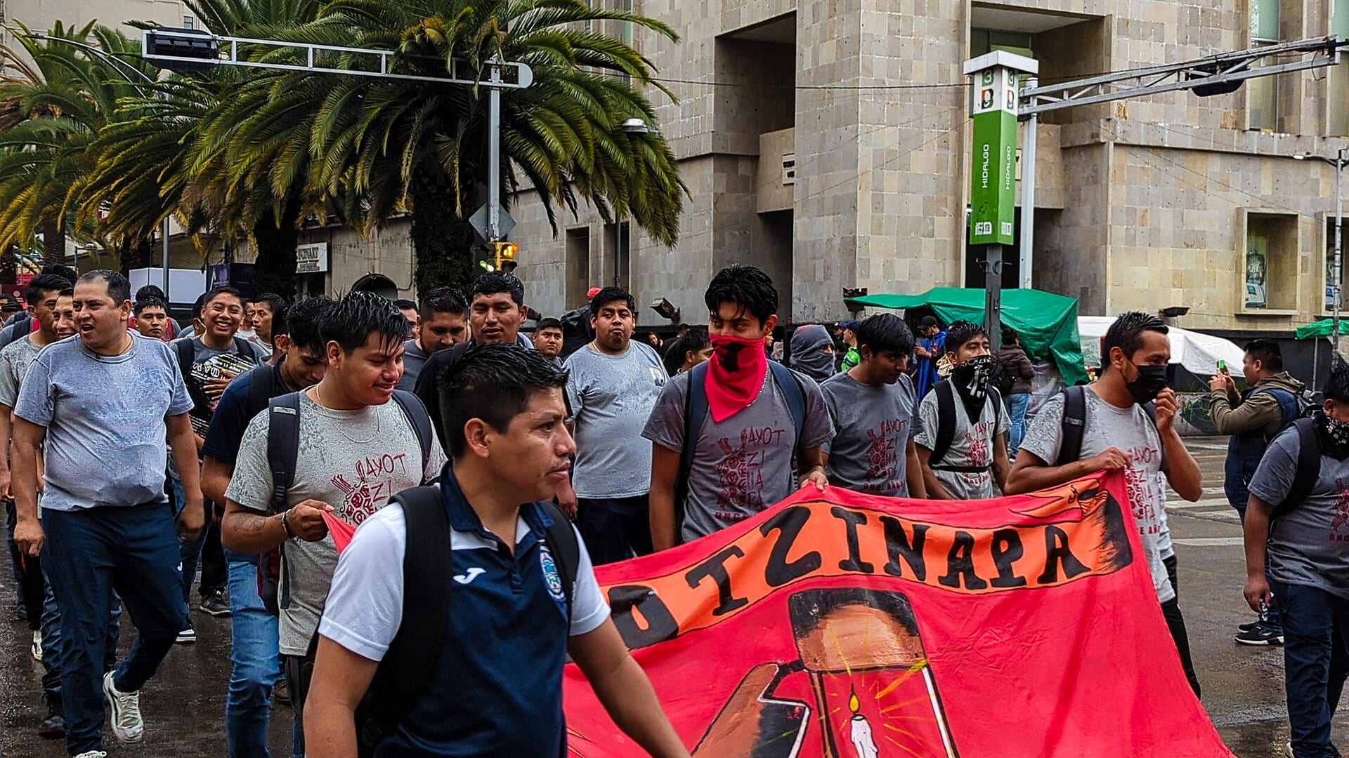 Jóvenes con playeras grises marchan por una calle mojada y sostienen una manta roja con la palabra Ayotzinapa.