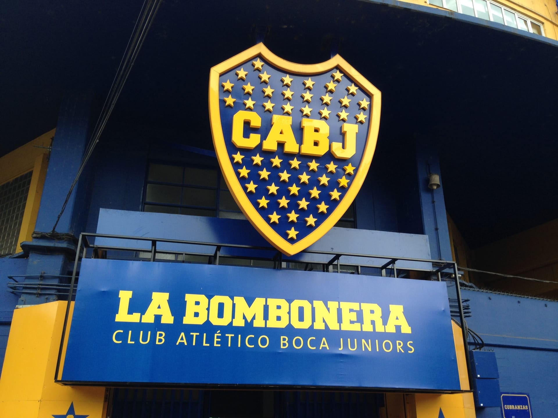 Escudo dorado y azul con las letras CABJ sobre un letrero que dice La Bombonera, en la fachada del estadio.