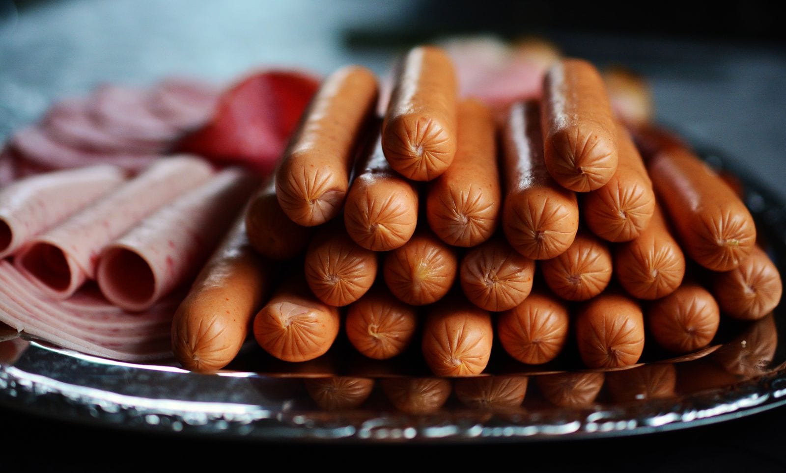 Una pila de salchichas tipo hot dog sobre una charola metálica, junto a rebanadas de jamón enrolladas.