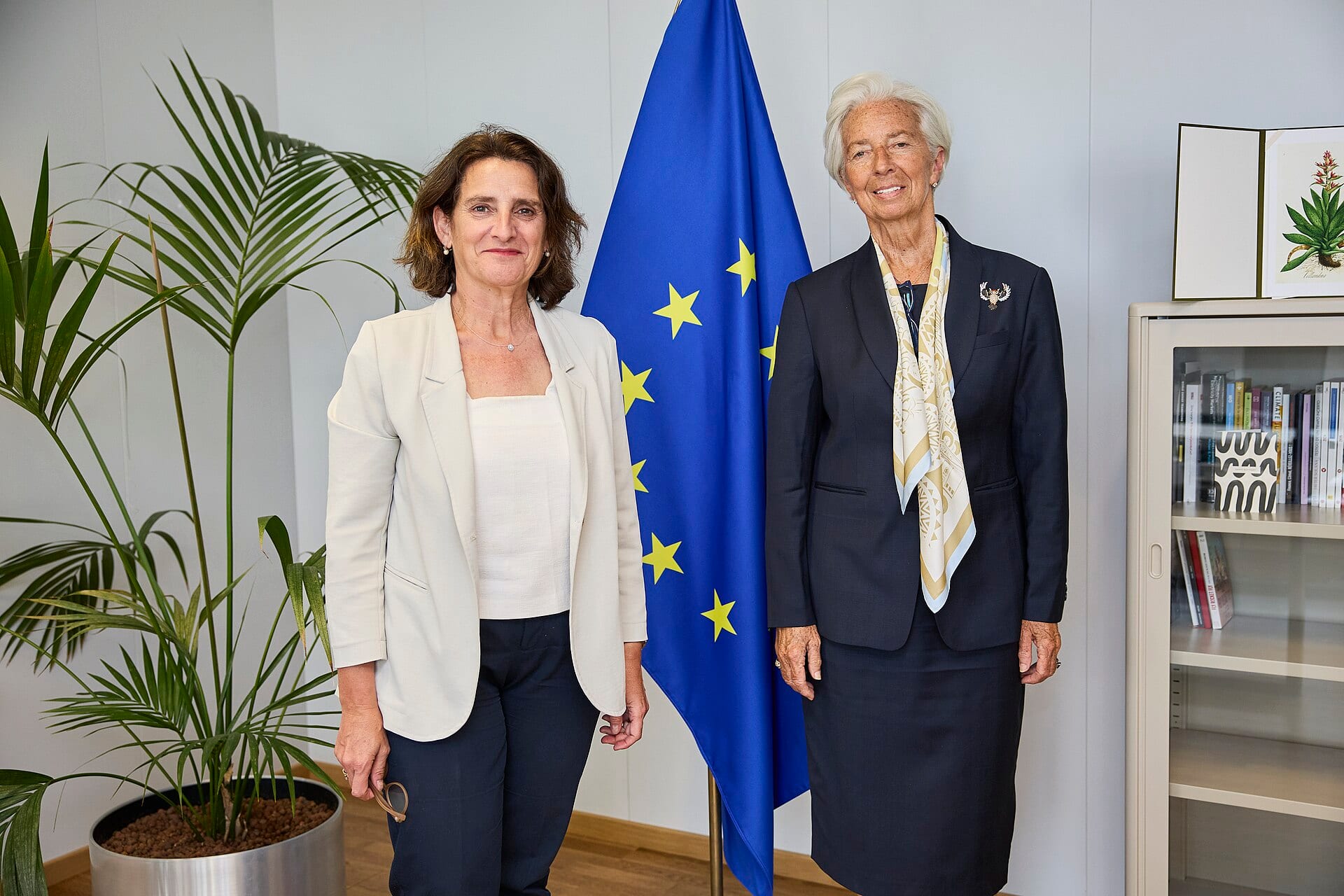 Dos mujeres posan de pie ante una bandera azul de la Unión Europea; Christine Lagarde, a la derecha, viste traje oscuro y pañuelo claro.