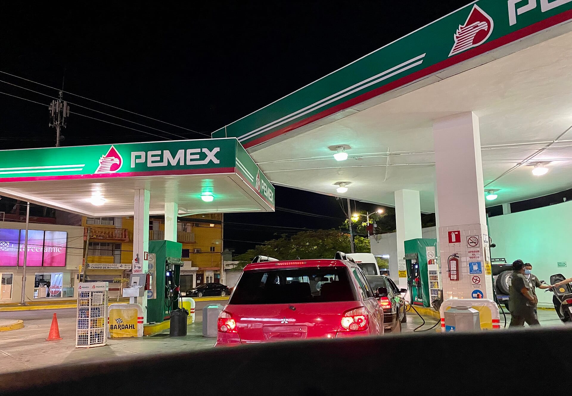 Una gasolinera de Pemex de noche, con techumbre verde y blanca iluminada y una camioneta roja esperando frente a los surtidores.