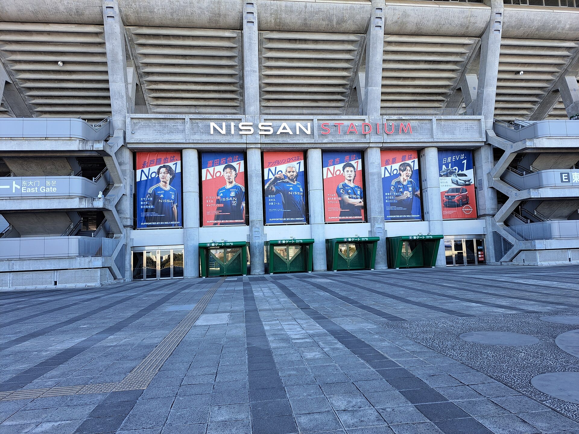 Fachada del estadio de Yokohama con el letrero Nissan Stadium y seis carteles de jugadores sobre las puertas del acceso este.