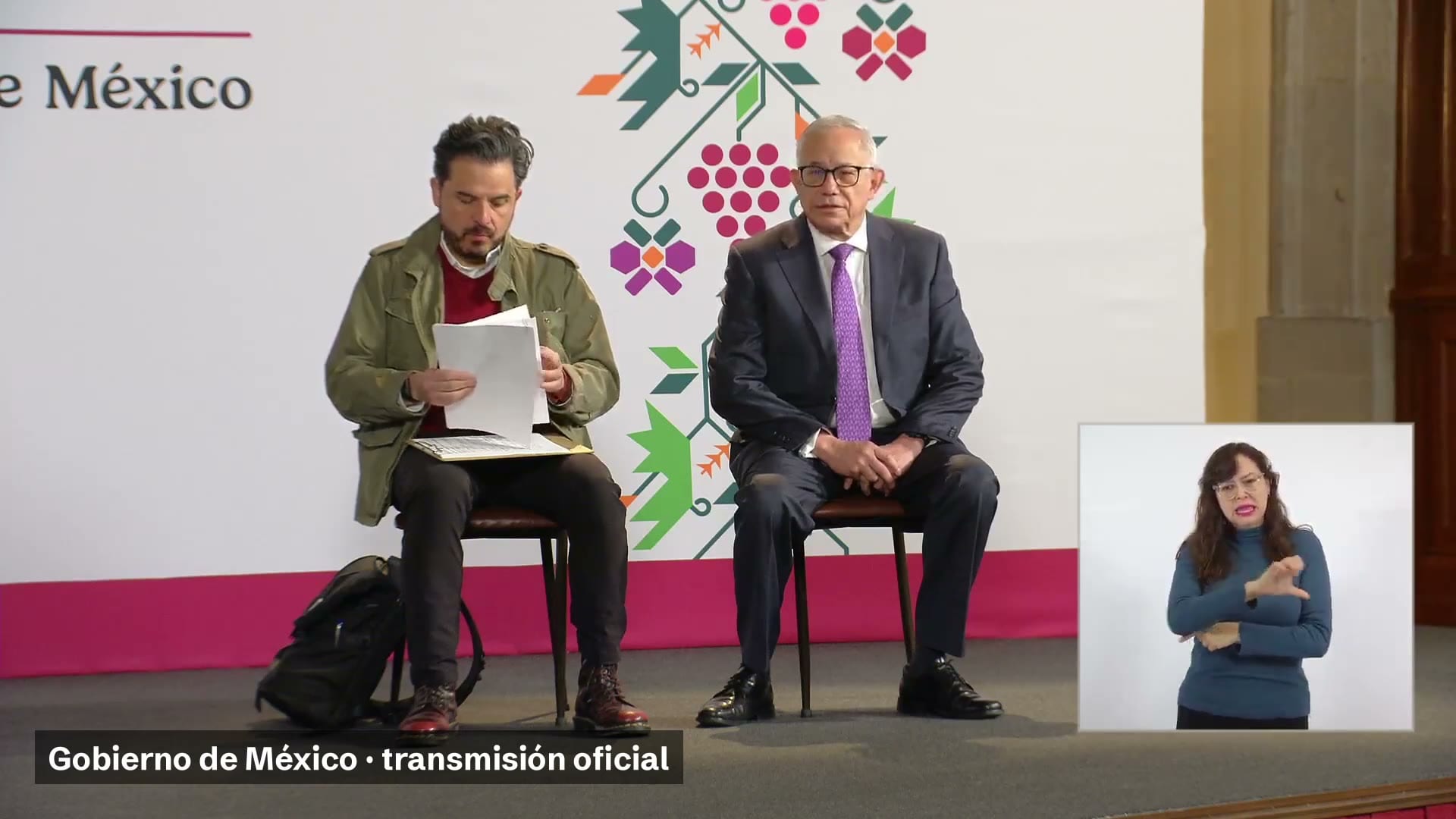 Dos hombres sentados en el estrado de la conferencia matutina; uno revisa unas hojas y, a la derecha, una intérprete de lengua de señas.