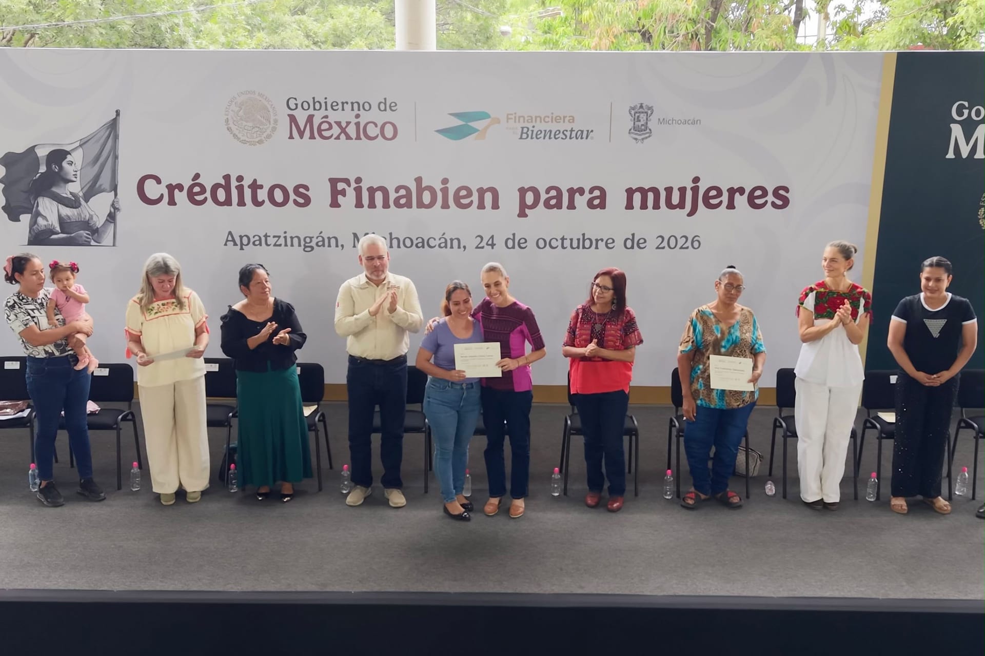 Claudia Sheinbaum abraza a una mujer que sostiene su constancia de crédito, frente a una lona que dice Créditos Finabien para mujeres, en Apatzingán.