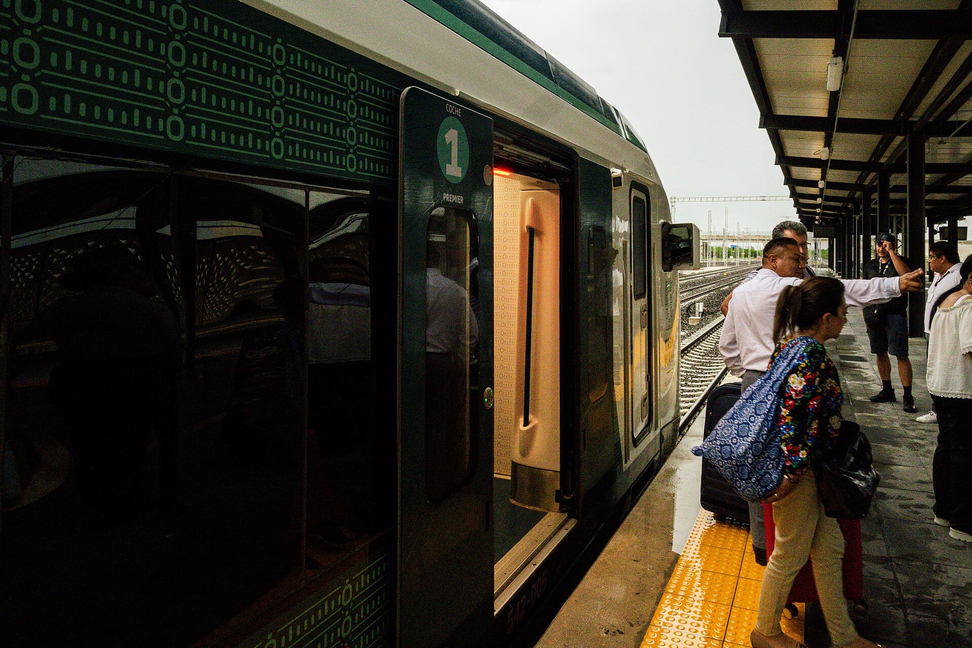 Pasajeros con maletas abordan por la puerta del coche 1 de un tren verde y blanco en un andén techado.