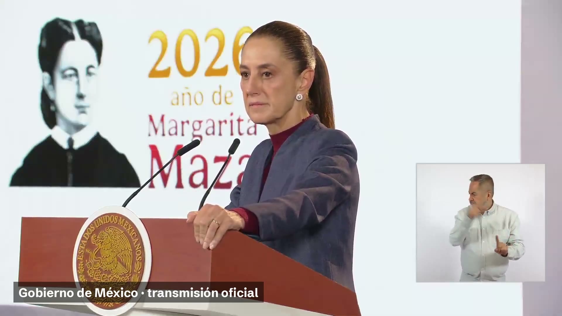 La presidenta Claudia Sheinbaum, de pie ante el podio con el escudo nacional, durante la conferencia matutina; detrás, una lámina del año de Margarita Maza.