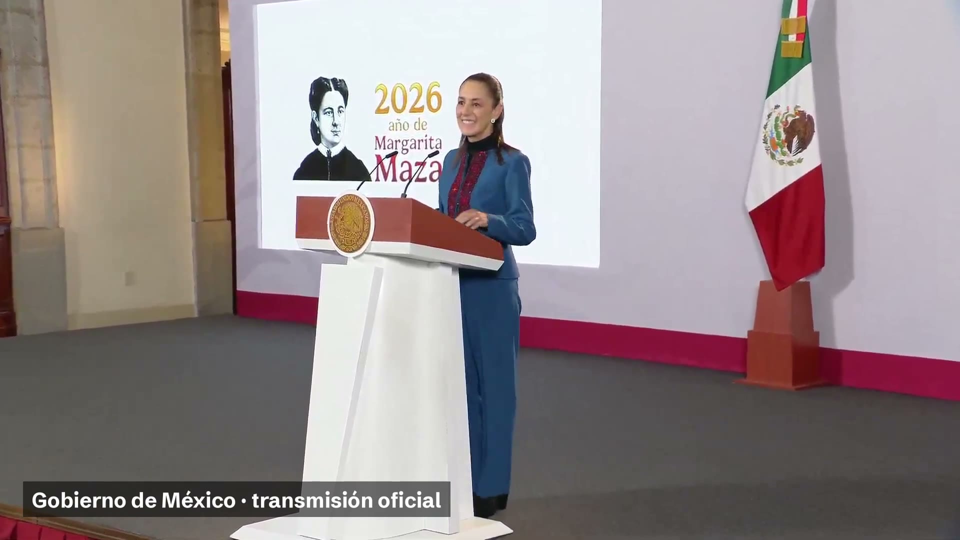 La presidenta Claudia Sheinbaum sonríe de pie frente a un podio, con una lámina del año de Margarita Maza detrás y una bandera de México a su derecha.