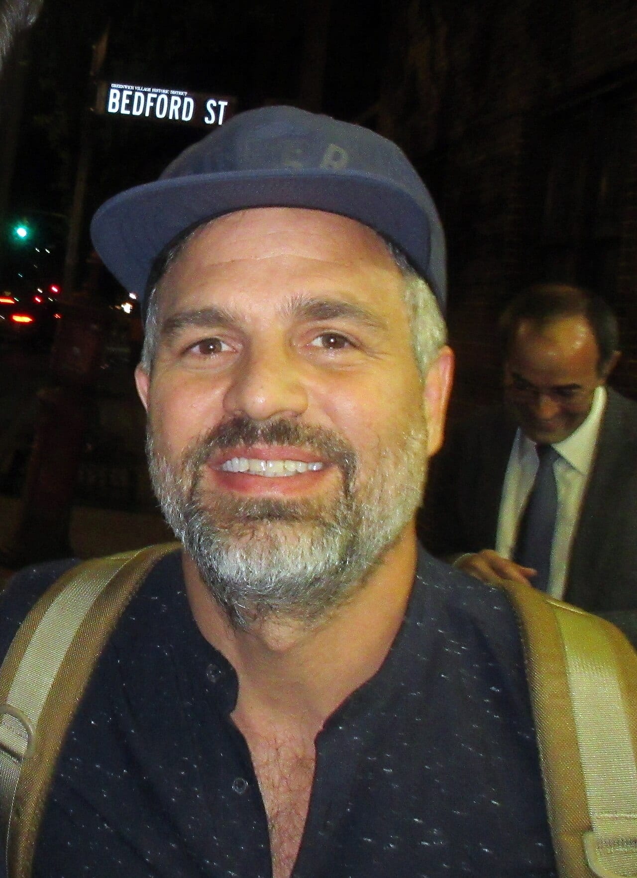 Un hombre con gorra azul, barba entrecana y mochila al hombro sonríe a la cámara de noche en una calle; detrás camina otro hombre de traje.