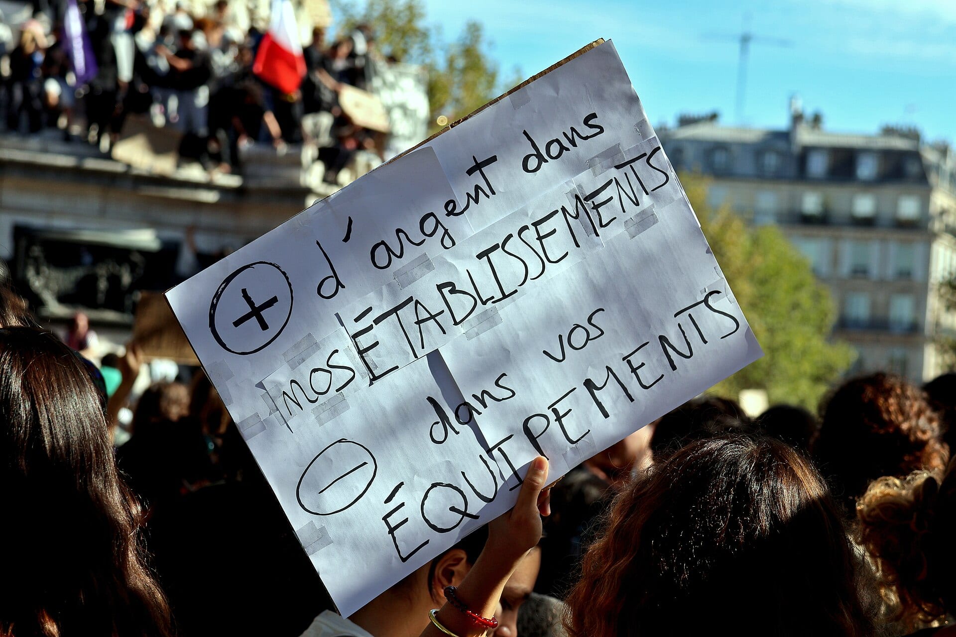 Una manifestante levanta un cartón en francés que pide más dinero para los establecimientos escolares y menos para los equipos, con una multitud detrás en una plaza de París.