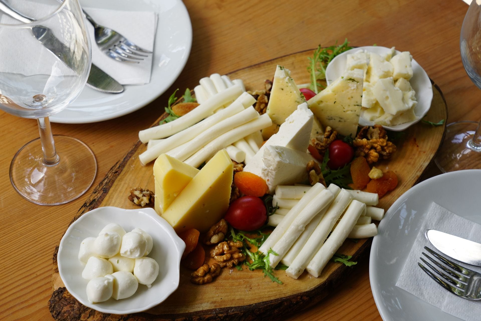 Tabla de madera con quesos blancos y amarillos, nueces, jitomates cherry y albaricoques secos, entre platos y copas.