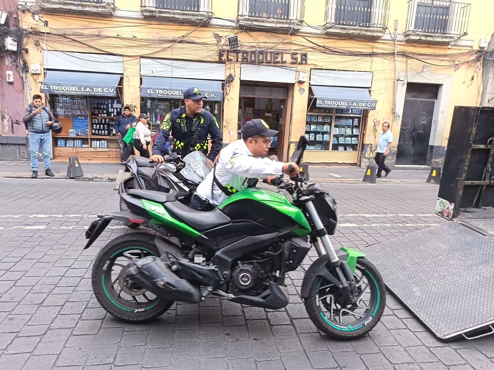 Un agente de tránsito empuja una motocicleta verde hacia la rampa de una grúa en una calle empedrada del Centro, frente a locales comerciales.