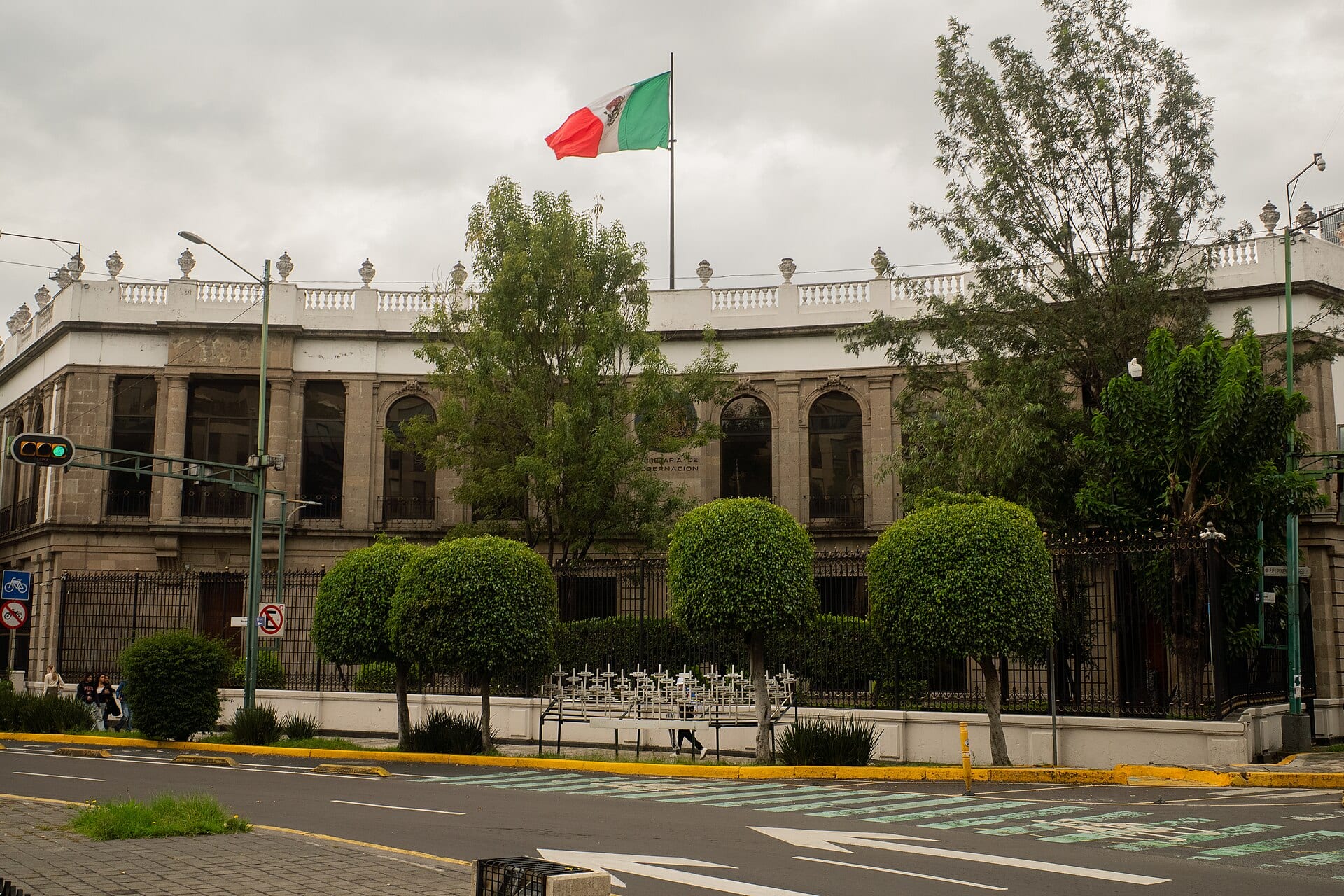 Fachada de piedra del Palacio de Cobián, con una bandera de México en el asta y árboles recortados frente a la reja.