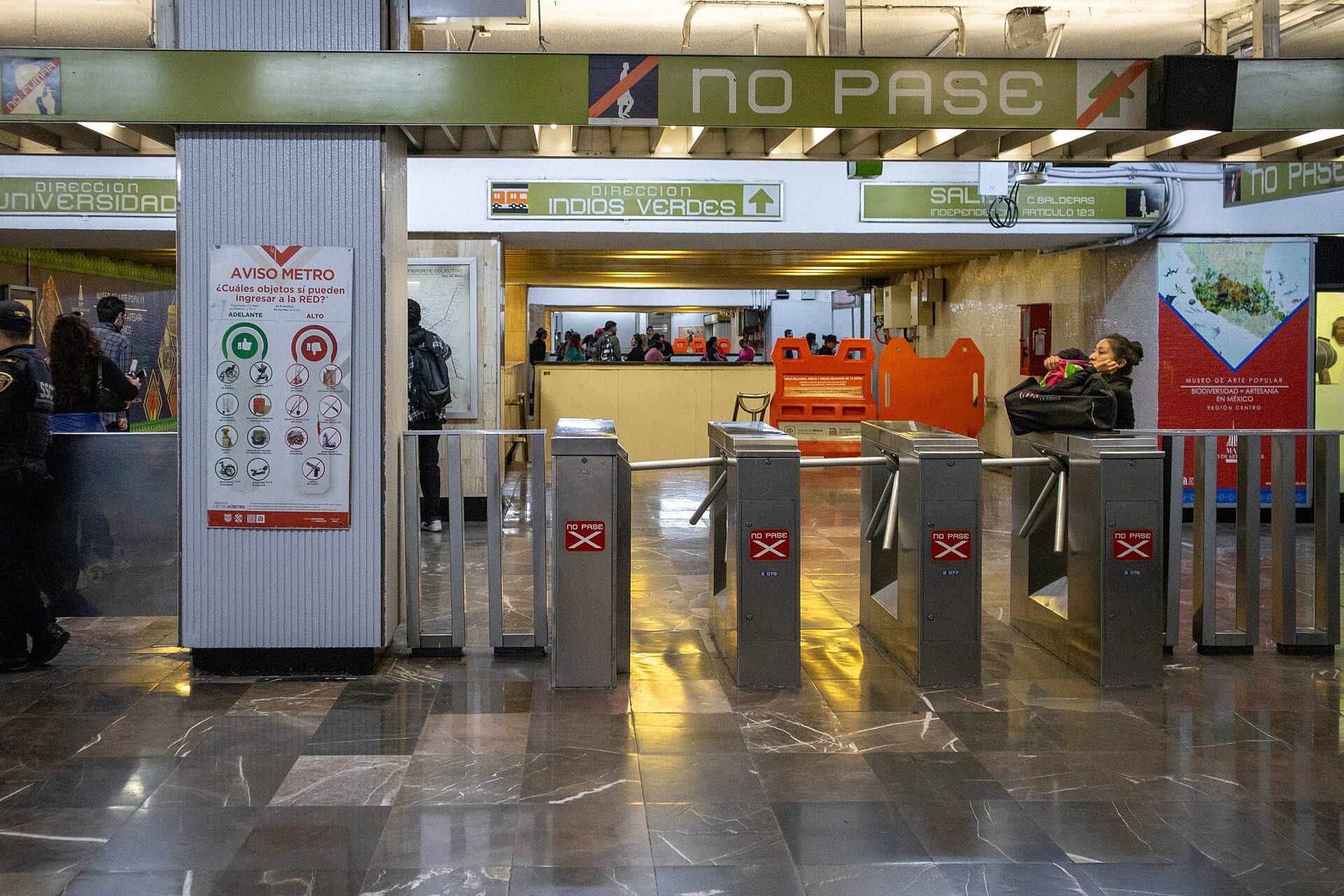 Torniquetes y letreros de una estación del Metro de la Ciudad de México, con un policía a la izquierda y usuarios al fondo.