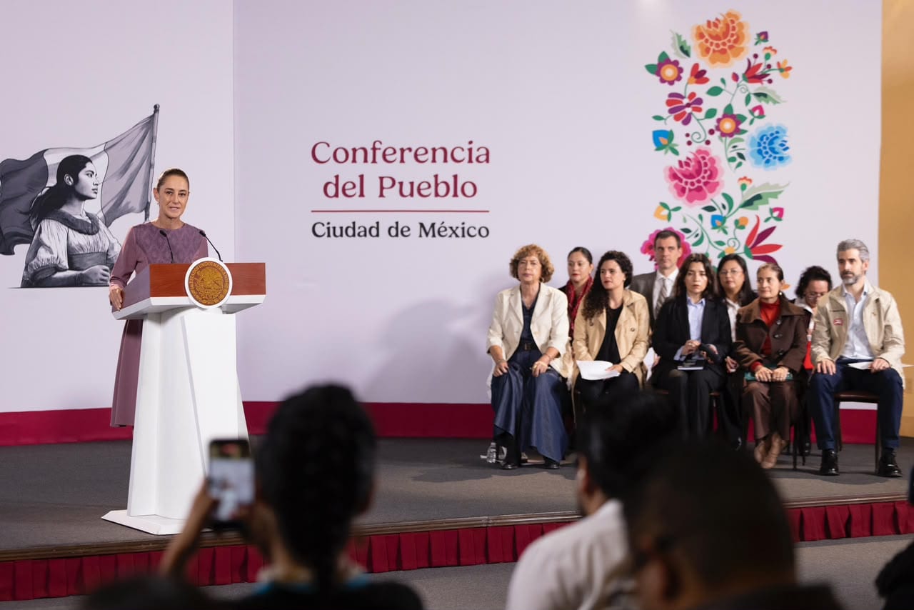 La presidenta Claudia Sheinbaum habla tras un podio con el escudo nacional, en un escenario con el letrero Conferencia del Pueblo, mientras varias personas sentadas la escuchan.
