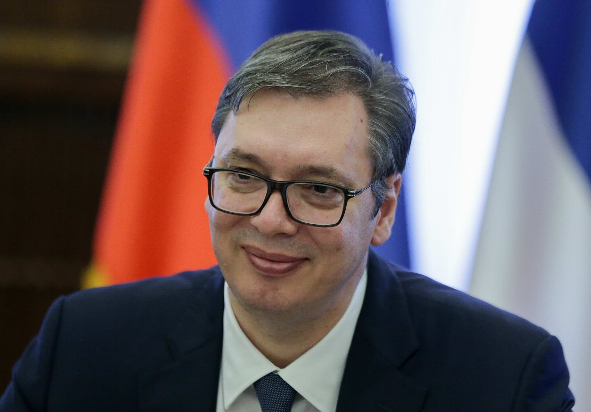 Aleksandar Vučić, con anteojos y traje oscuro, sonríe frente a unas banderas de colores.