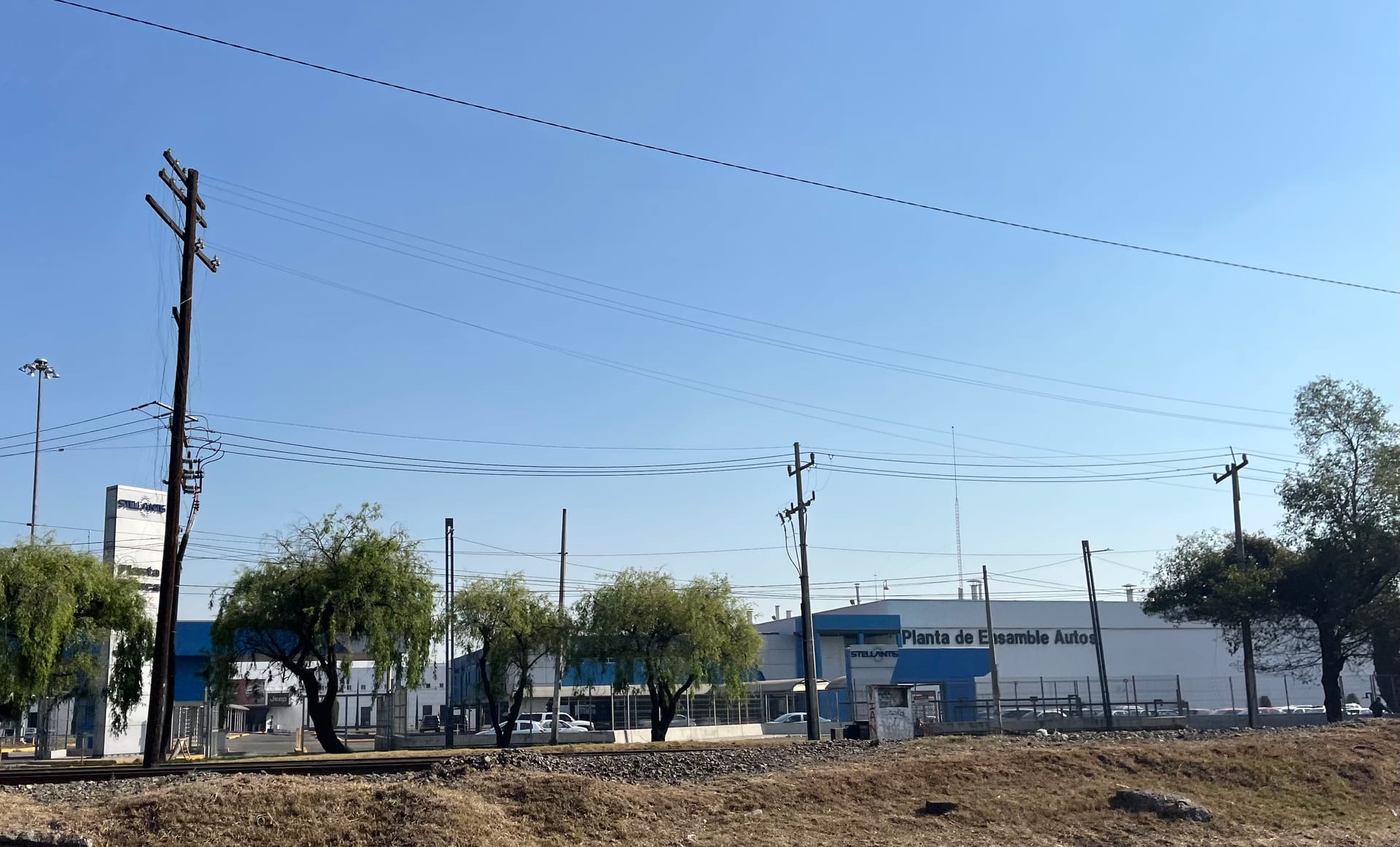 The front of the Stellantis car assembly plant in Toluca, with trees, utility poles and a clear sky.