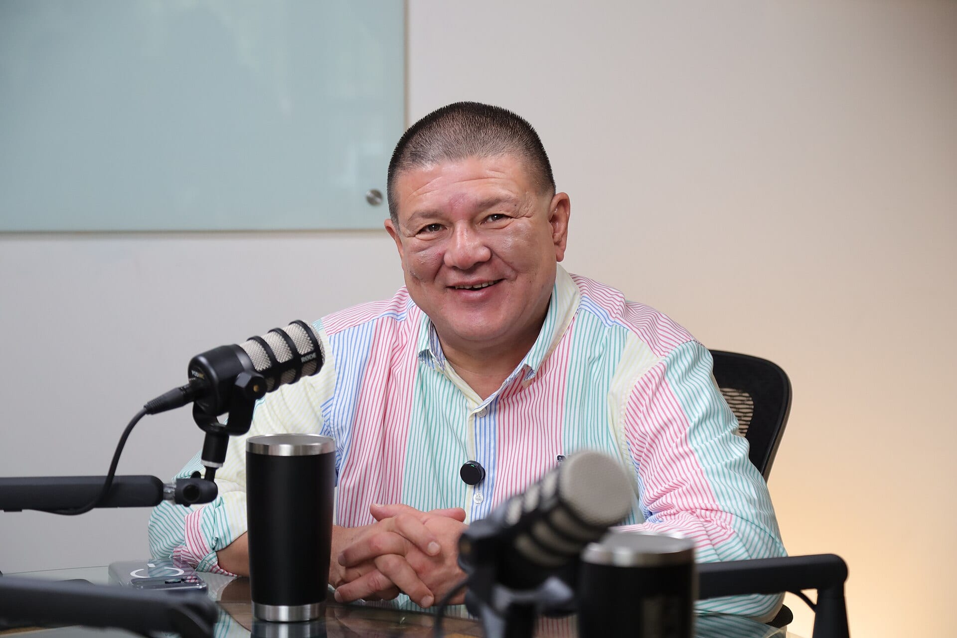 Un hombre de pelo corto y camisa de rayas de colores sonríe sentado ante dos micrófonos de estudio.