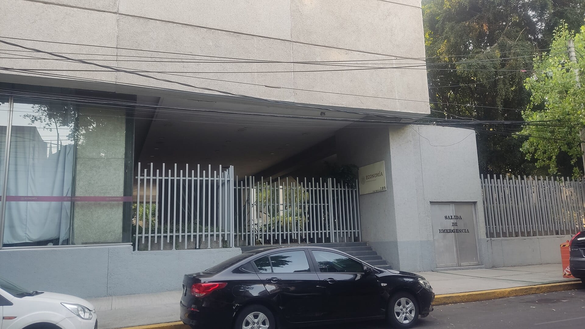 Entrada de un edificio de la Secretaría de Economía, con un letrero del escudo y el nombre de la dependencia junto a una reja blanca.