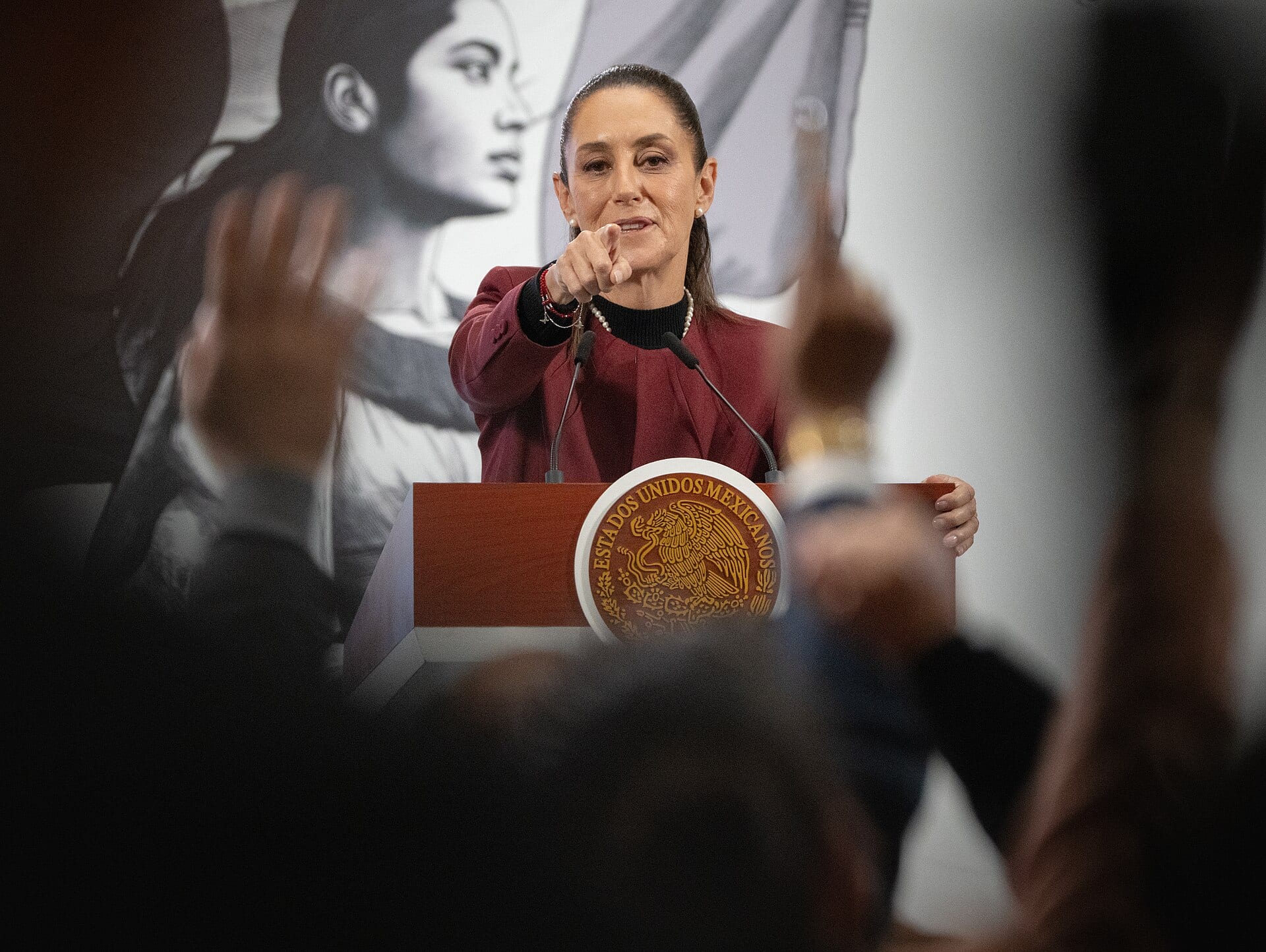 Una mujer de saco guinda señala hacia el público desde un podio con el escudo nacional, entre manos levantadas.