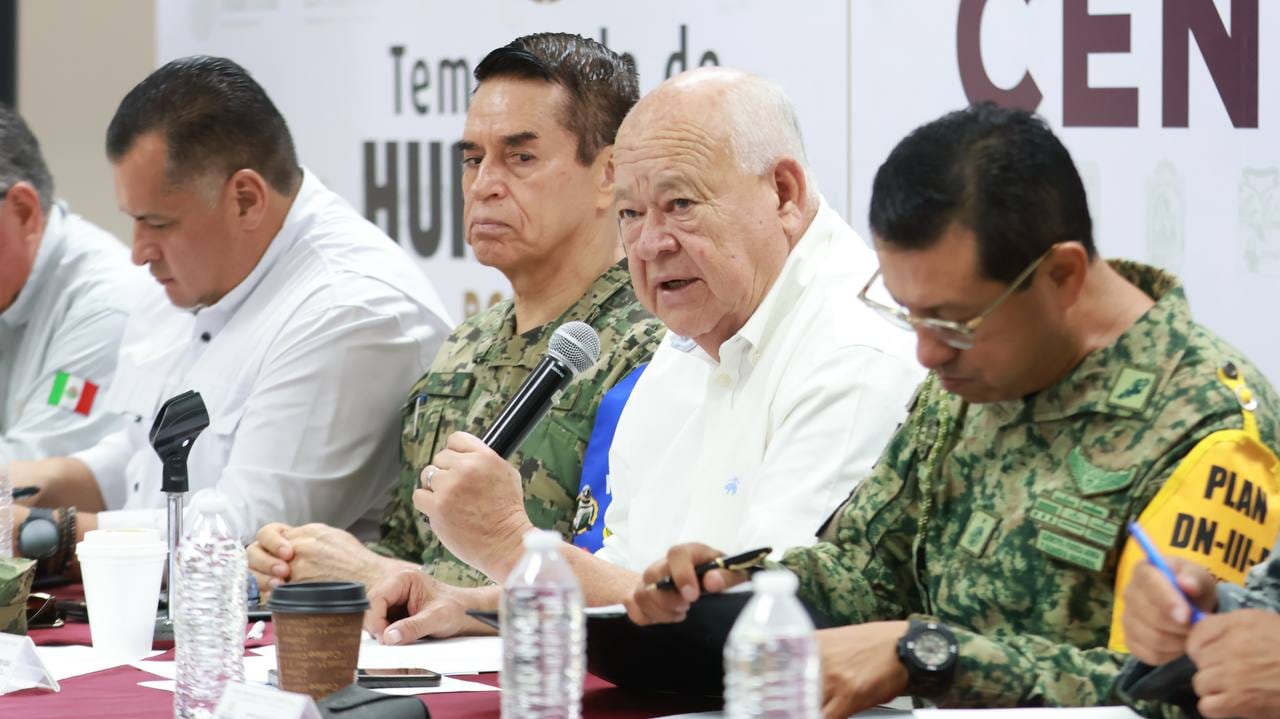 Un funcionario habla al micrófono en una mesa junto a mandos navales y del Ejército, con un cartel sobre la temporada de huracanes al fondo.