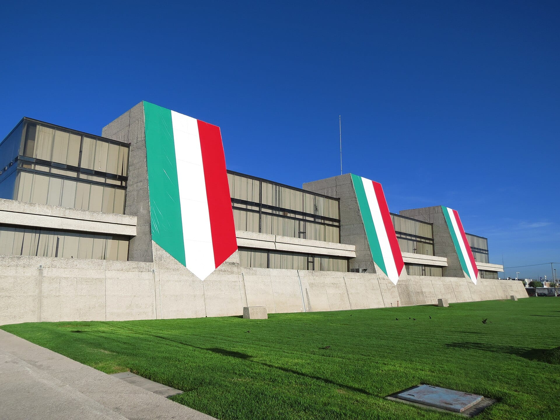 Edificio de concreto y vidrio del INEGI con tres banderas de México colgadas en sus torres, bajo un cielo azul.