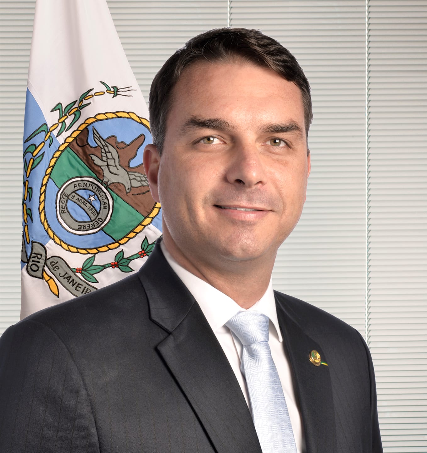 Portrait of Senator Flávio Bolsonaro in a dark suit and light tie, beside the flag of the state of Rio de Janeiro.
