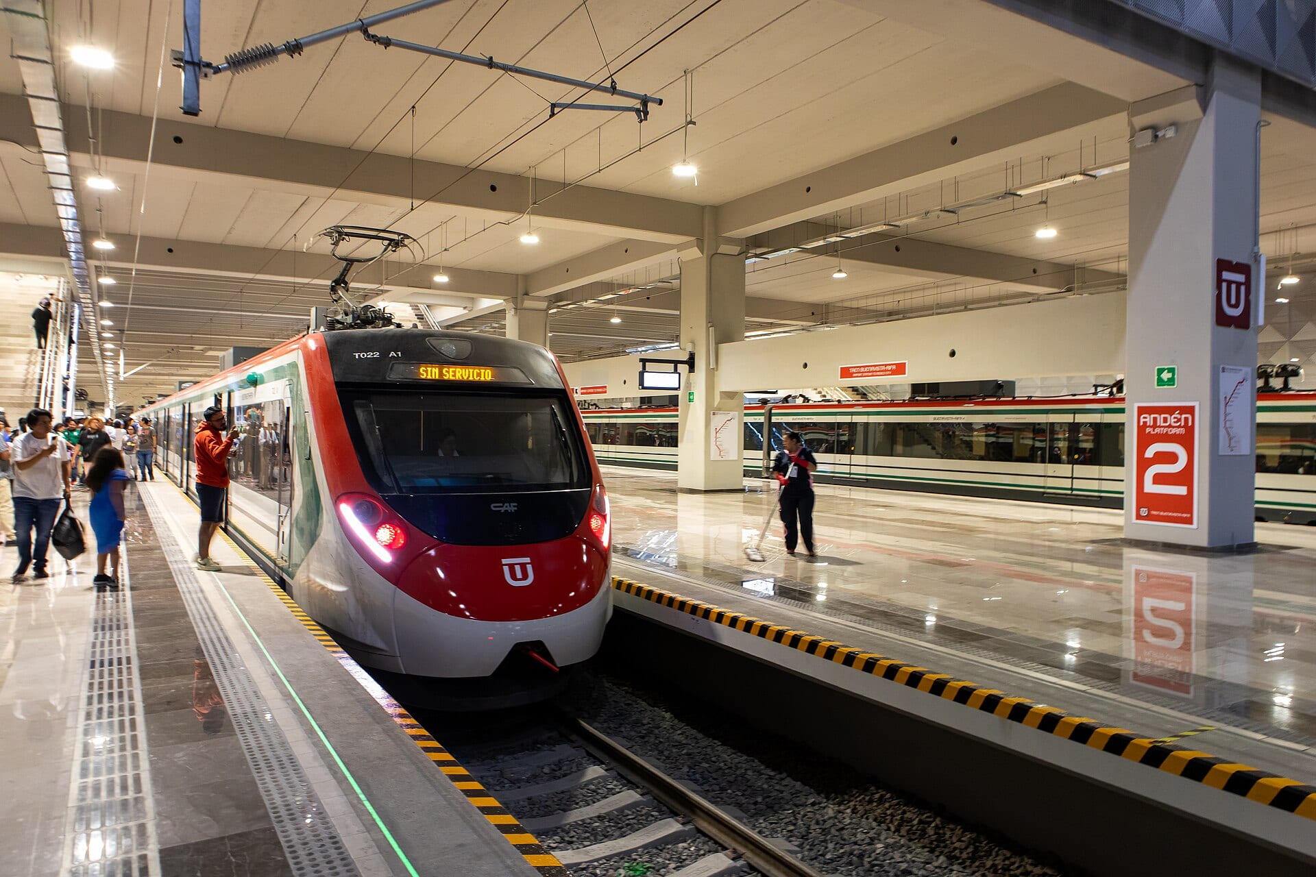 Un tren con frente rojo y blanco y el letrero Sin servicio se detiene en un andén, con pasajeros a la izquierda y otro tren al fondo.