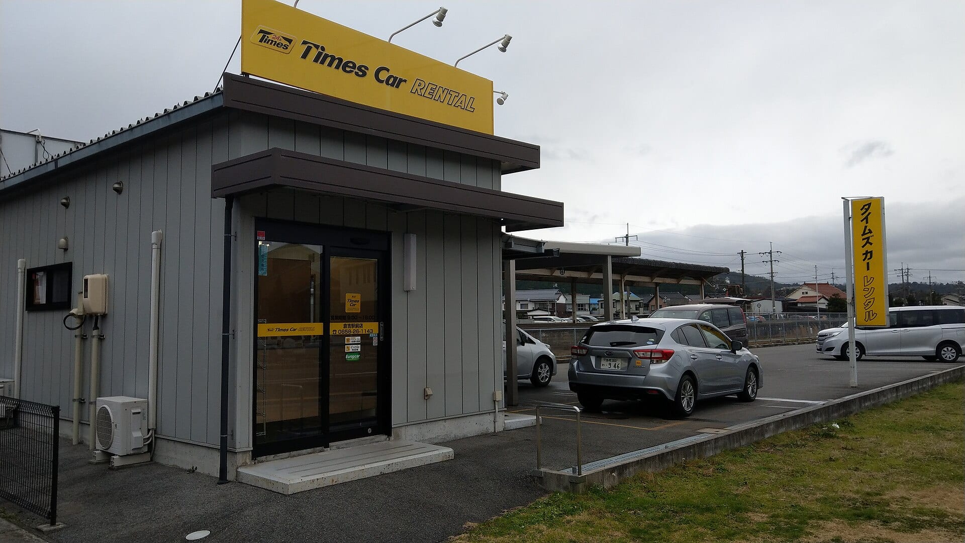 Oficina de renta de autos con un letrero amarillo de Times Car Rental, un automóvil gris estacionado y un cielo nublado.