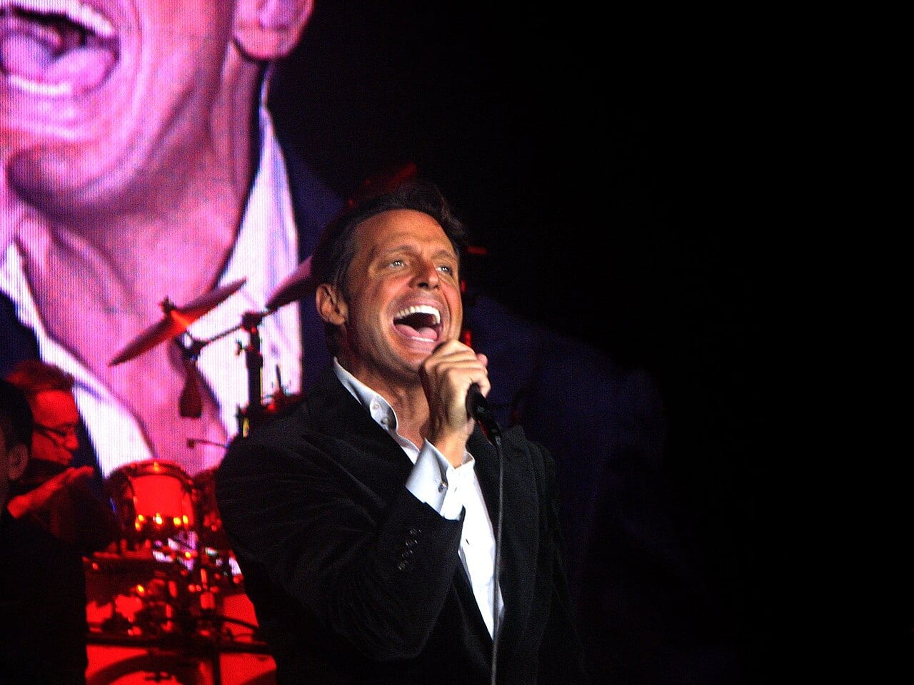 Luis Miguel canta frente a un micrófono sobre un escenario, con un traje oscuro y luces detrás.