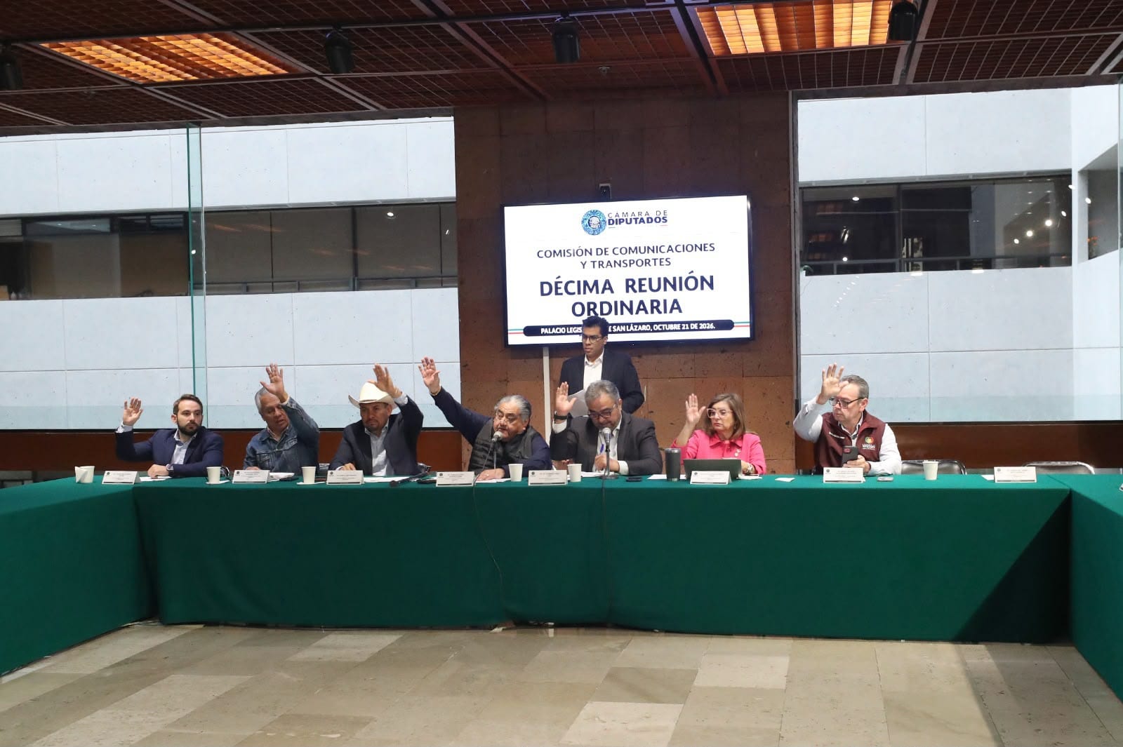 Siete integrantes de la Comisión de Comunicaciones y Transportes levantan la mano para votar en una mesa con mantel verde, bajo una pantalla que anuncia su décima reunión ordinaria.
