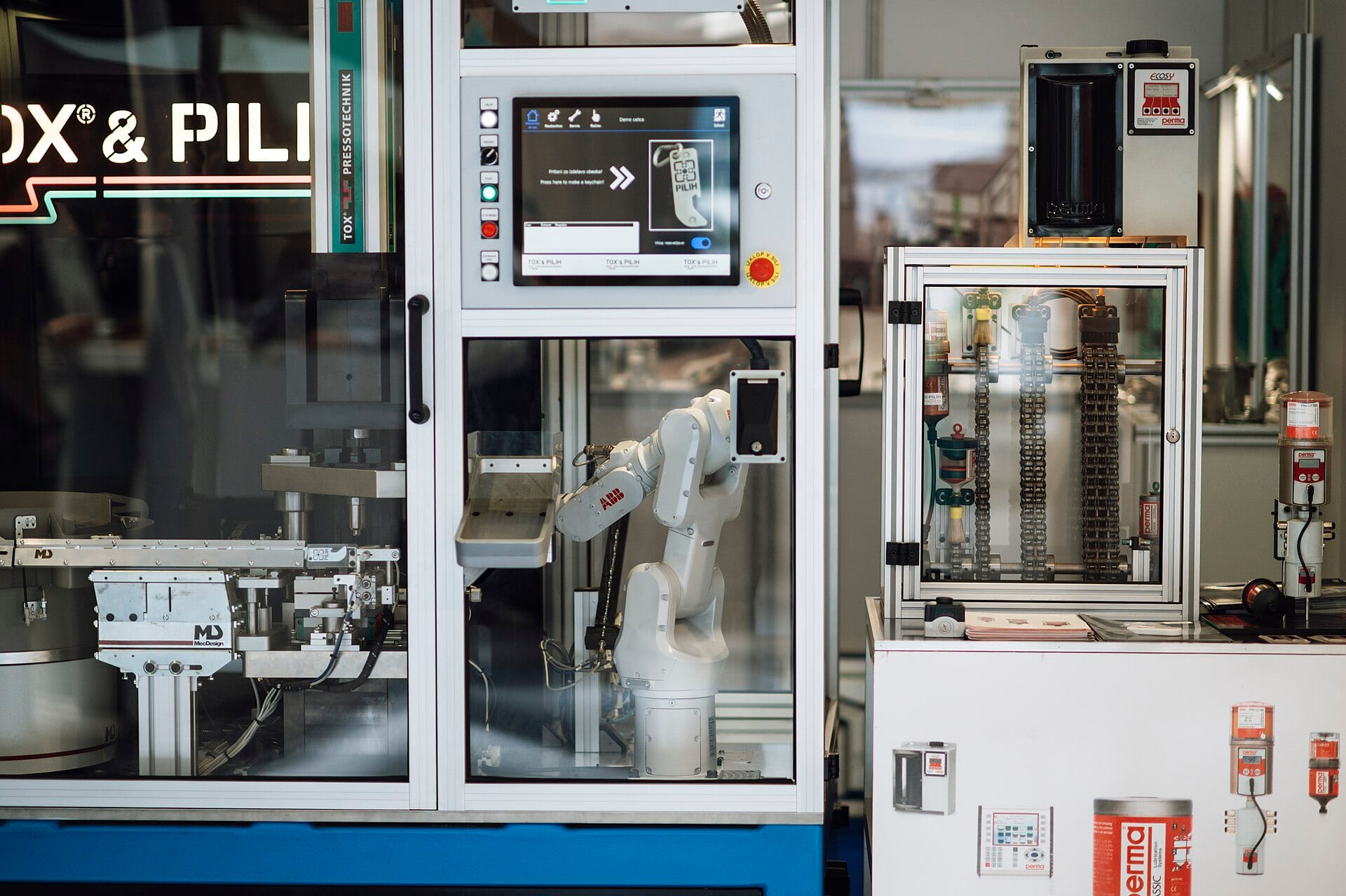A white robotic arm works inside a glass cell next to a control screen at an industrial fair.