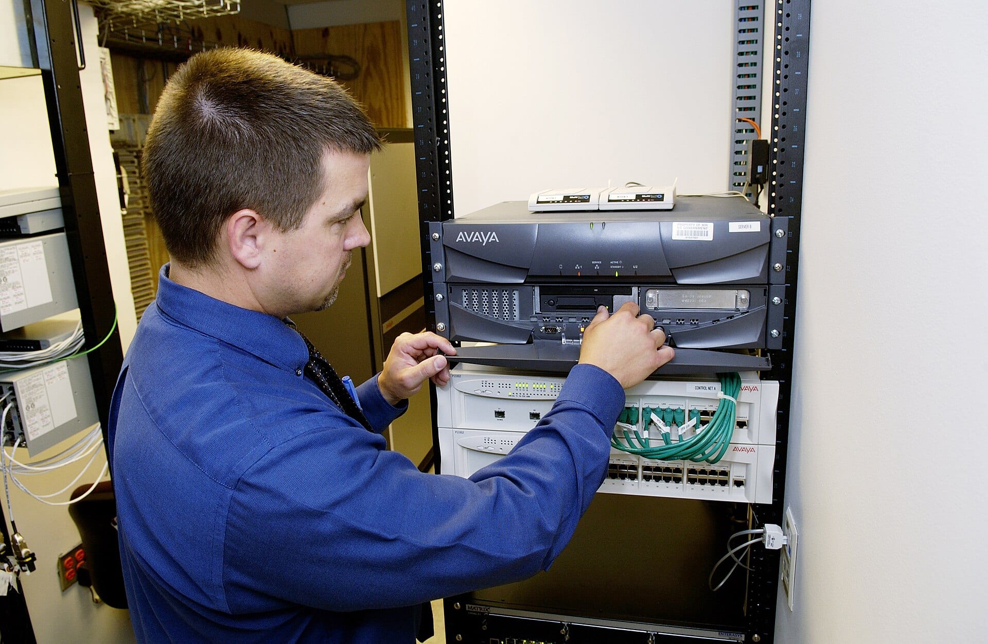A technician in a blue shirt checks a server and a network switch mounted in a rack.