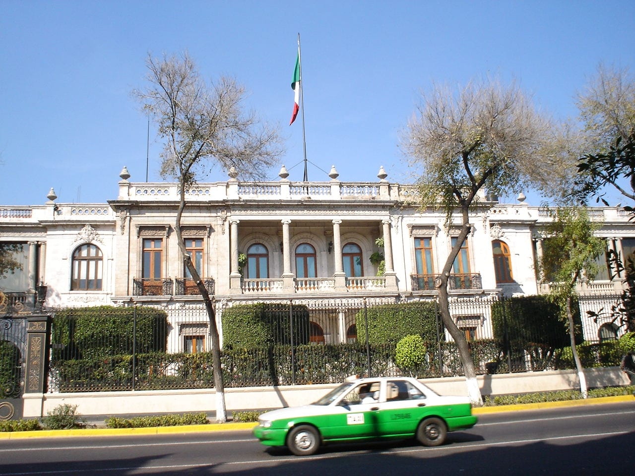 Fachada de un edificio de dos pisos con balcón y columnas, bandera de México en el techo y un taxi verde y blanco pasando frente a la reja.