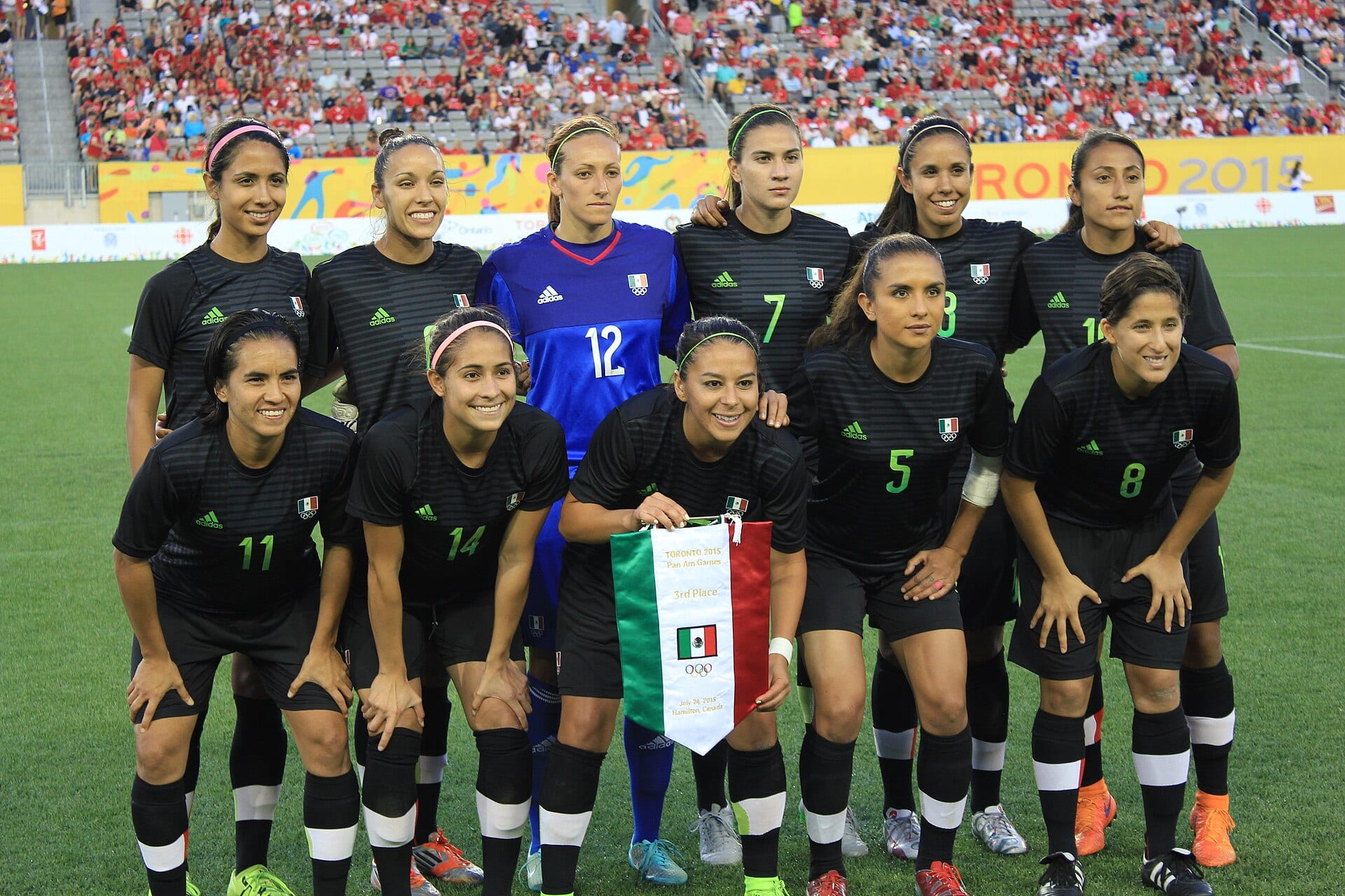 Once jugadoras de México con uniforme negro posan en dos filas sobre el césped, con una portera de azul al centro y un banderín tricolor frente a ellas.